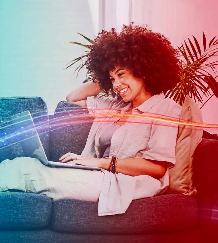 A imagem mostra uma mulher sentada no sofá e navegando na internet com o notebook no colo. 
